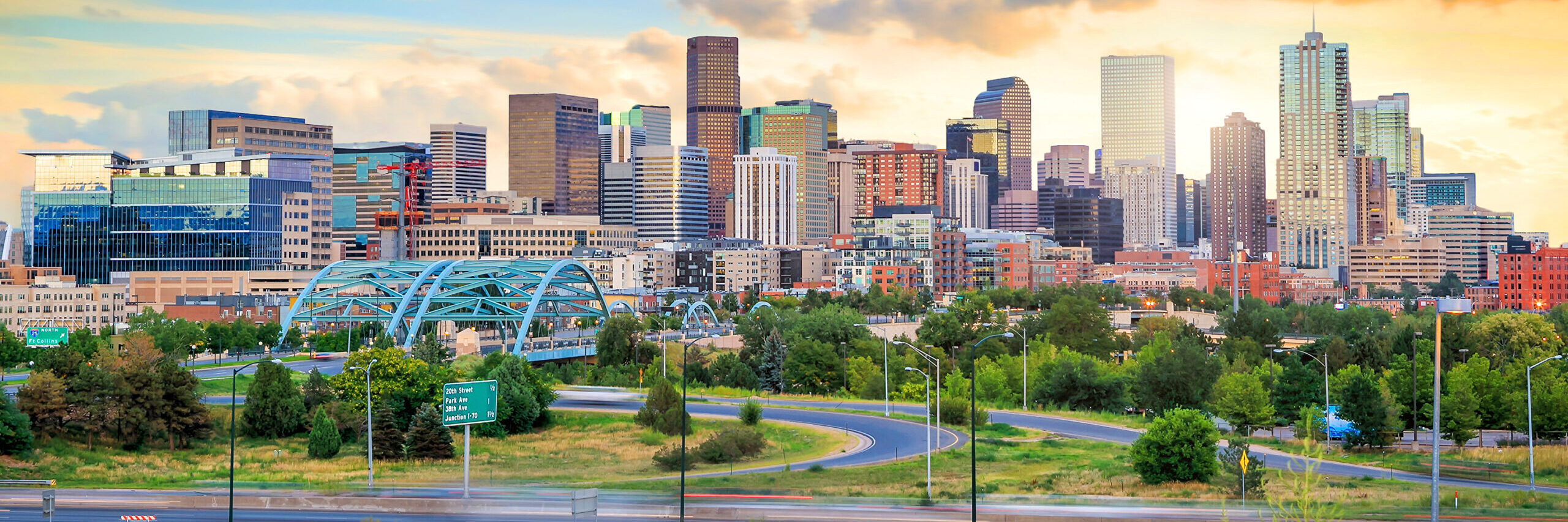 Denver, Colorado skyline with downtown buildings, trees, and a roadway in the foreground.