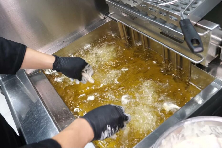 Gloved hands drop battered pieces into a large commercial deep fryer full of bubbling golden oil, with a fry basket raised on the right and a bowl of battered food to the lower right.