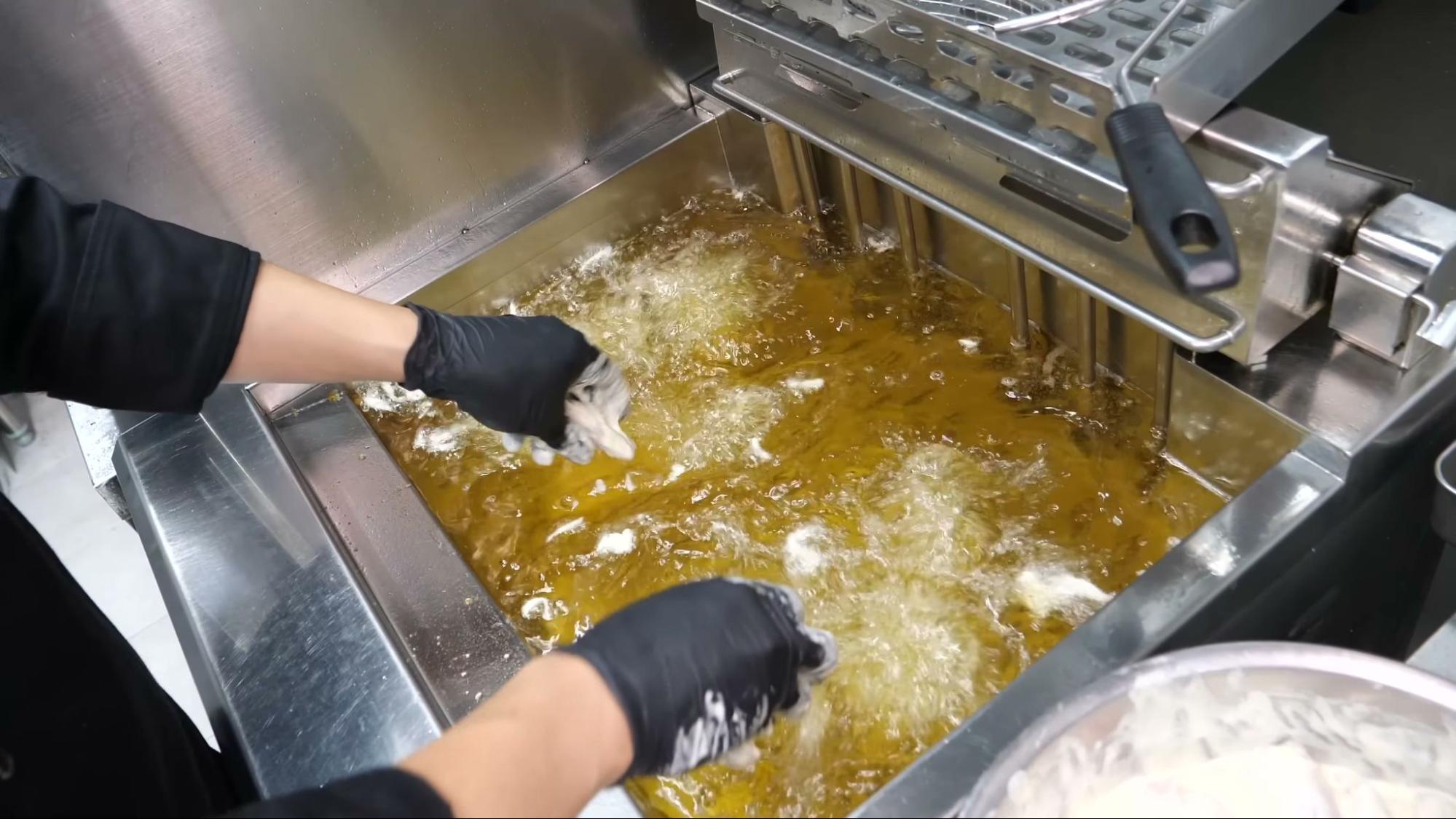 Gloved hands drop battered pieces into a large commercial deep fryer full of bubbling golden oil, with a fry basket raised on the right and a bowl of battered food to the lower right.