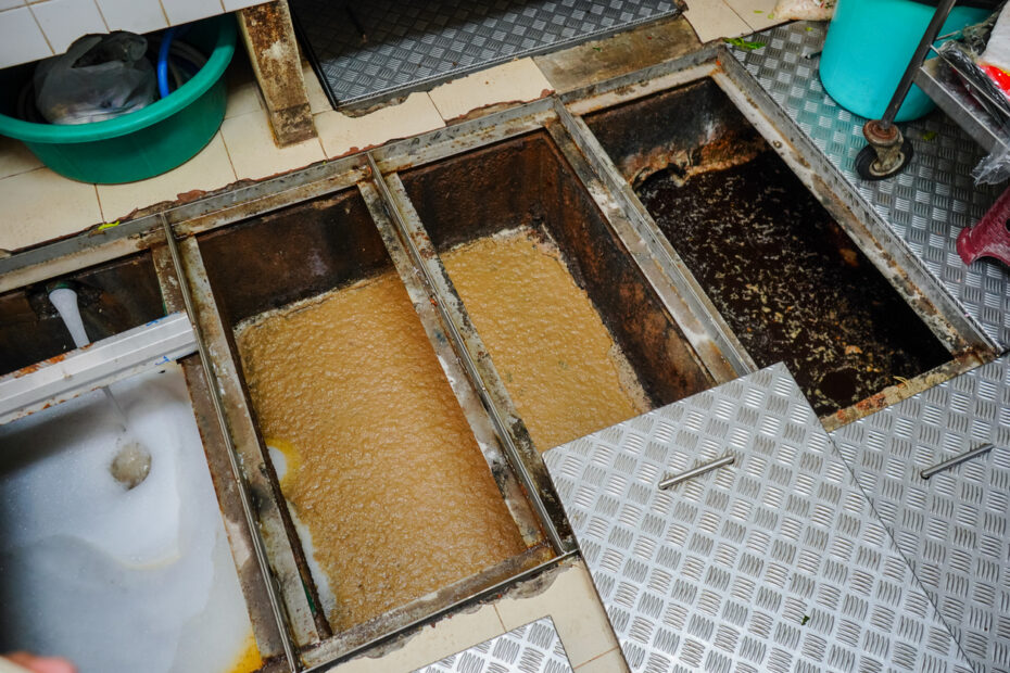 An open in-floor grease trap in a commercial kitchen, its metal diamond-plate covers set aside, showing multiple compartments filled with brown greasy wastewater and accumulated fats.