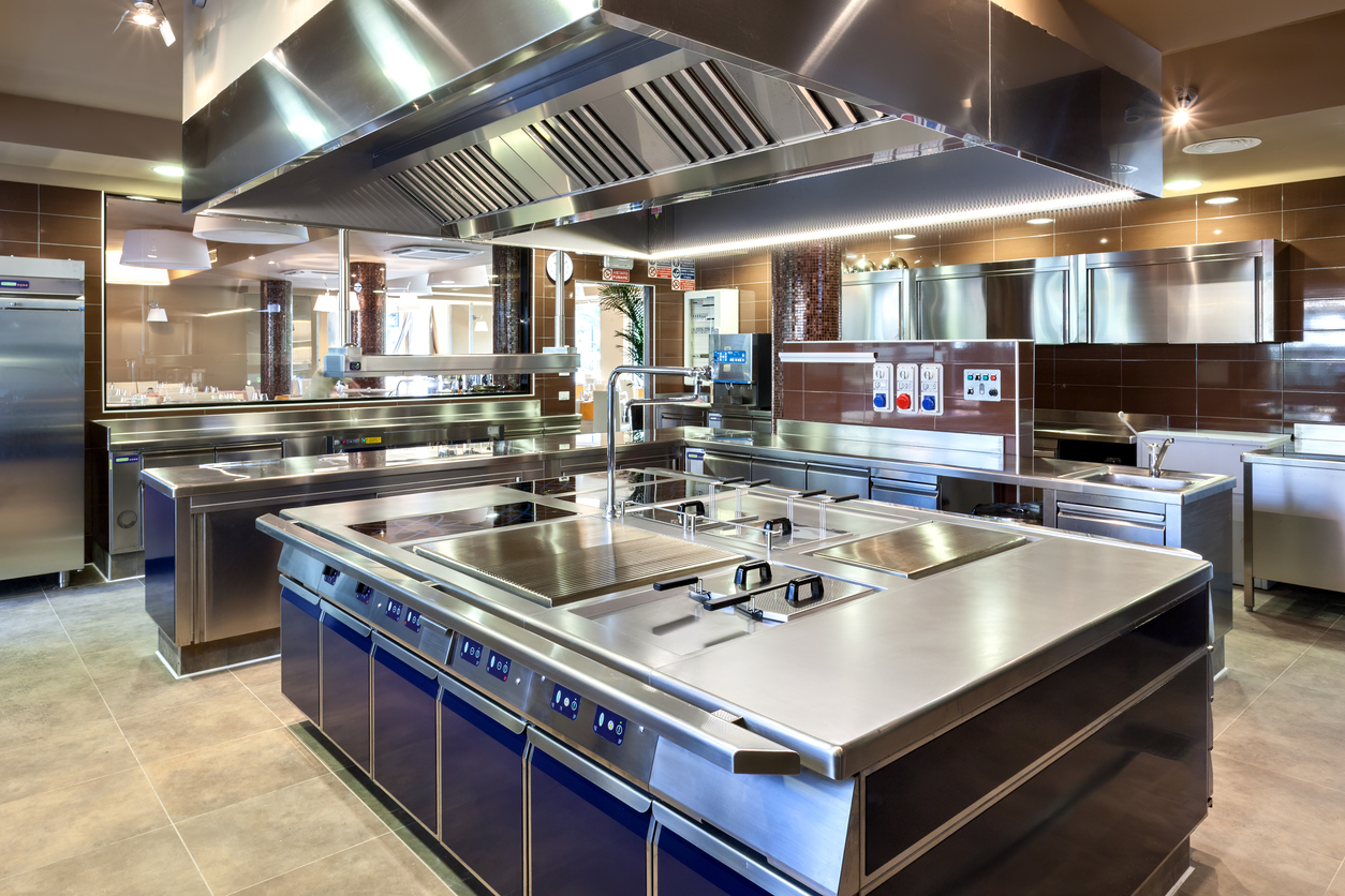 Modern commercial kitchen with stainless steel cooking equipment, large ventilation hood, preparation surfaces, and food service workstations.