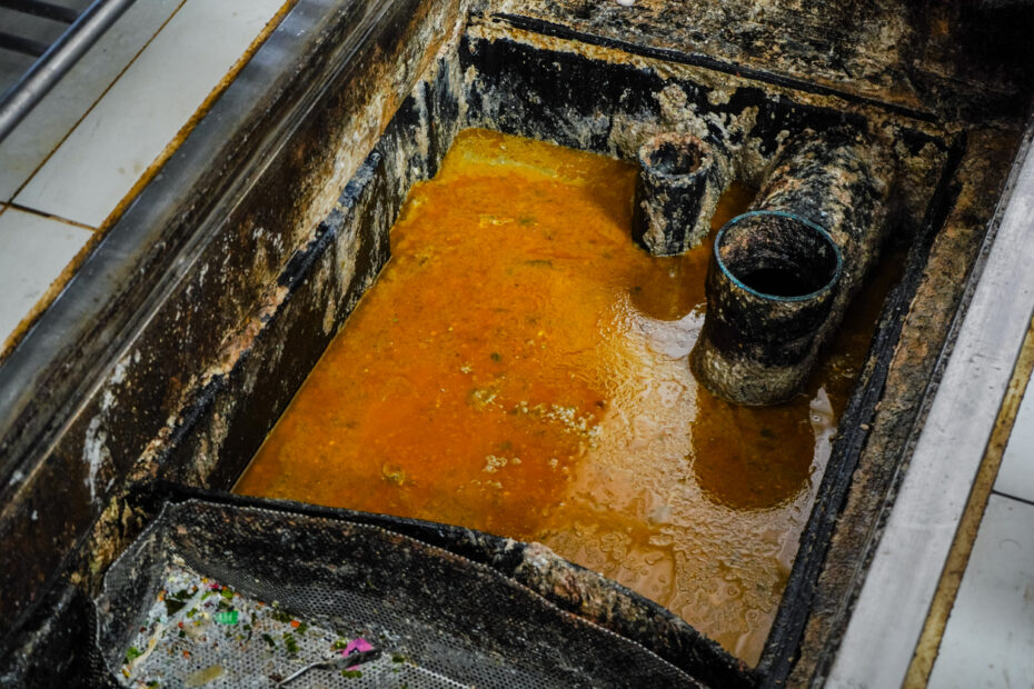Dirty grease trap containing standing wastewater and buildup, showing heavy grease residue and grime around the interior walls and pipes.