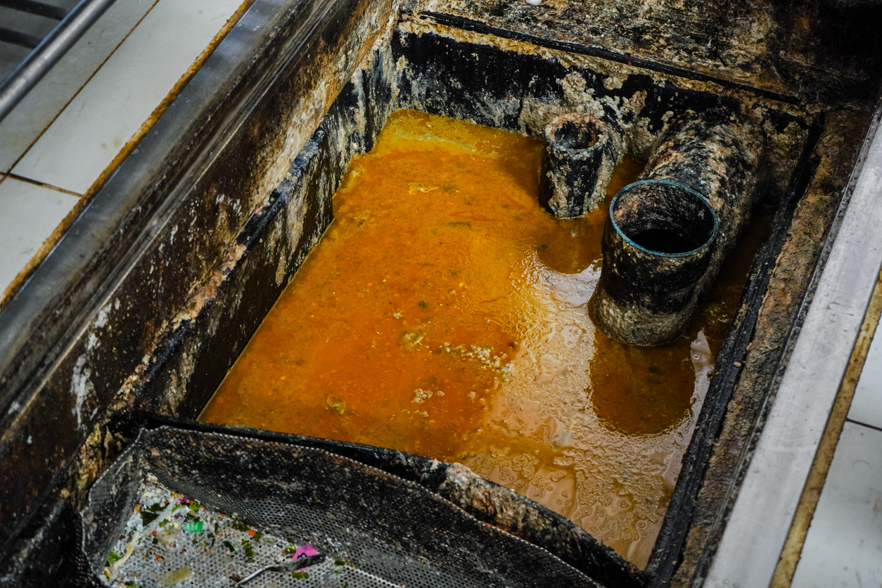 Dirty grease trap containing standing wastewater and buildup, showing heavy grease residue and grime around the interior walls and pipes.