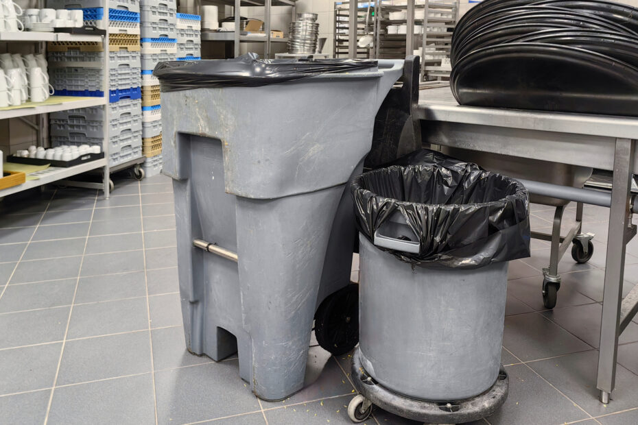 Commercial kitchen waste bins and trash cans positioned beside stainless steel prep tables and dish racks.