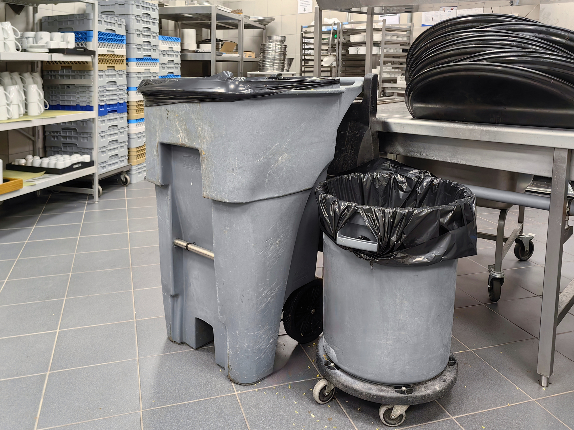Commercial kitchen waste bins and trash cans positioned beside stainless steel prep tables and dish racks.